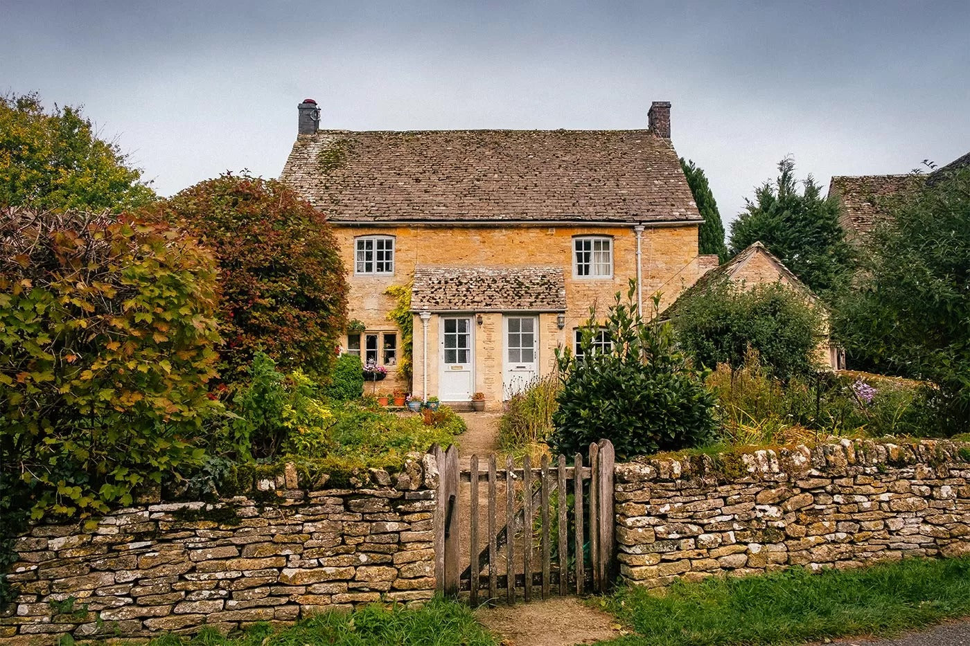 Charming honey-stone cottage in the Cotswolds with a slate roof, lush garden, and weathered stone wall with wooden gate in the foreground.