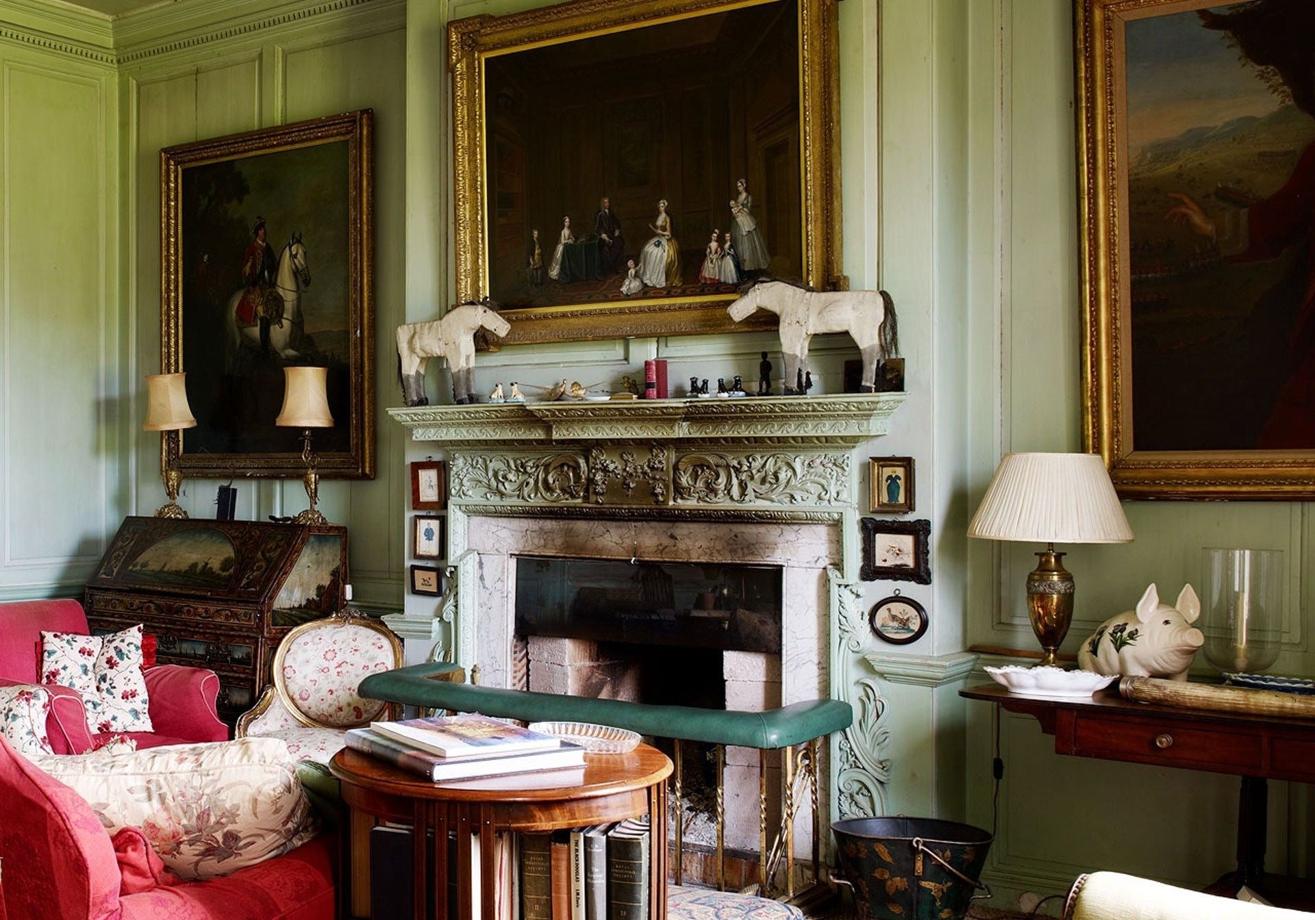 Traditional English country drawing room with green panelled walls, gilded portraits, antique fireplace, and layered textiles.
