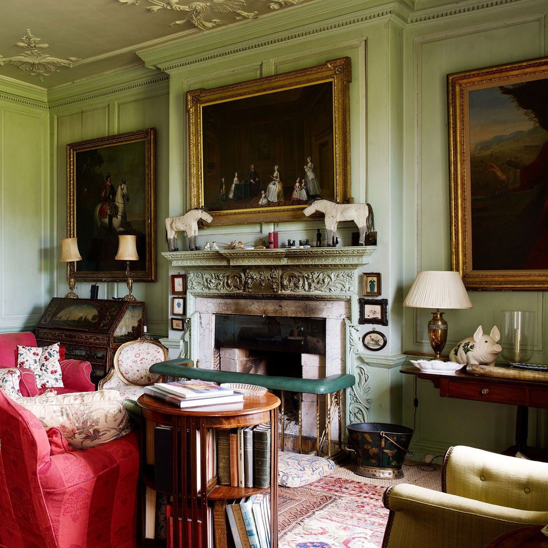 Traditional English country drawing room with green panelled walls, gilded portraits, antique fireplace, and layered textiles.